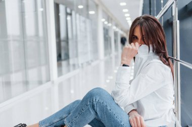 Beyaz gömlekli, elinde telefon olan esmer bir kadın modern havaalanında ya da koridorda gündüz vakti oturuyor..