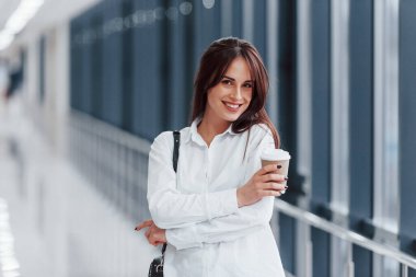 Modern havaalanında ya da koridorda elinde bir bardak içkiyle duran beyaz gömlekli esmer kadın..