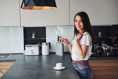 Genç, güzel esmer, günlük kıyafetlerle gündüzleri mutfakta akıllı telefon kullanıyor..