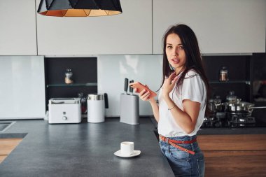 Genç, güzel esmer, günlük kıyafetlerle gündüzleri mutfakta akıllı telefon kullanıyor..