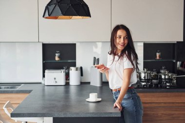Genç, güzel esmer, günlük kıyafetlerle gündüzleri mutfakta akıllı telefon kullanıyor..