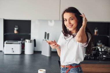 Genç, güzel esmer, günlük kıyafetlerle gündüzleri mutfakta akıllı telefon kullanıyor..
