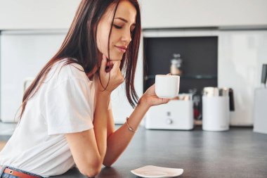 Gündelik giysiler içinde genç, güzel esmer sabah taze içeceklerle mutfakta oturuyor..