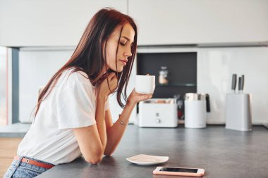 Gündelik giysiler içinde genç, güzel esmer sabah taze içeceklerle mutfakta oturuyor..