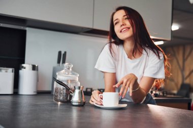 With cup of fresh coffee. Young beautiful brunette in casual clothes indoors in kitchen at daytime.