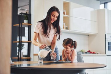 Mother pouring fresh milk into the glass for her little daughter in kitchen.