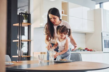 Mother pouring fresh milk into the glass for her little daughter in kitchen.