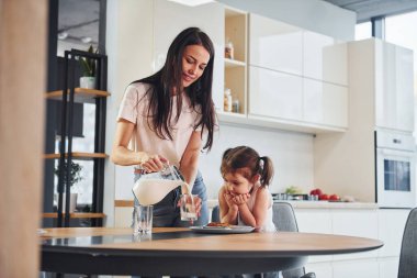 Mother pouring fresh milk into the glass for her little daughter in kitchen.