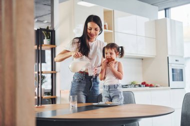Mother pouring fresh milk into the glass for her little daughter in kitchen.
