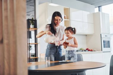 Mother pouring fresh milk into the glass for her little daughter in kitchen.