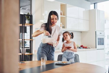 Mother pouring fresh milk into the glass for her little daughter in kitchen.