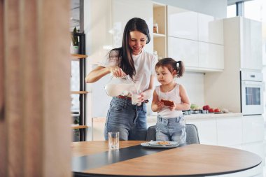 Mother pouring fresh milk into the glass for her little daughter in kitchen.
