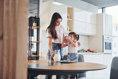 Mother pouring fresh milk into the glass for her little daughter in kitchen.