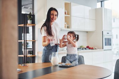 Mother pouring fresh milk into the glass for her little daughter in kitchen.