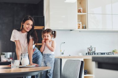 Little girl eats cookies. Mother with her little daughter indoors in kitchen.