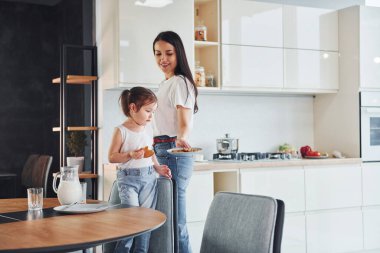 Little girl eats cookies. Mother with her little daughter indoors in kitchen.