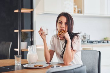 Genç ve güzel esmer, gündüz kıyafetleri içinde, mutfakta bir bardak süt ve şekerle..