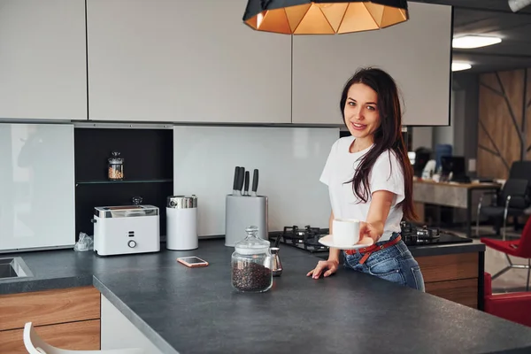 Gives fresh coffee. Young beautiful brunette in casual clothes indoors in kitchen at daytime.
