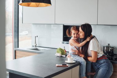 Mother with her little daughter embracing each other indoors in kitchen.