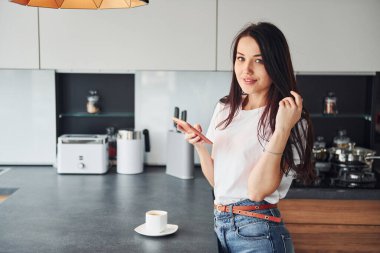 Genç, güzel esmer, günlük kıyafetlerle gündüzleri mutfakta akıllı telefon kullanıyor..