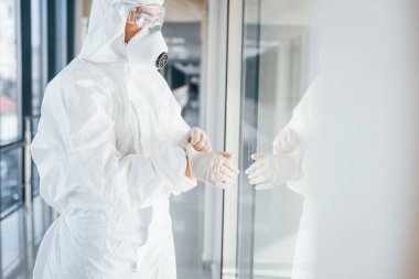 Laboratuvar önlüklü, koruyucu gözlüklü ve maskeli kadın doktor içeride duruyor ve eldiven giyiyor..