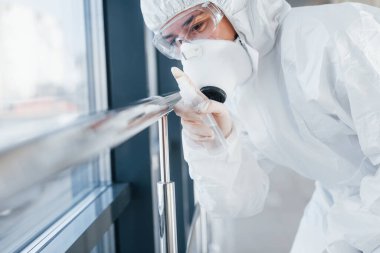 Female doctor scientist in lab coat, defensive eyewear and mask standing indoors with antibacterial spray.