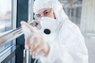 Laboratuvar önlüklü, koruyucu gözlüklü ve maskeli kadın doktor içeride antibakteriyel spreyle duruyor..