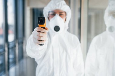 Female doctor scientist in lab coat, defensive eyewear and mask standing indoors with infrated thermometer.
