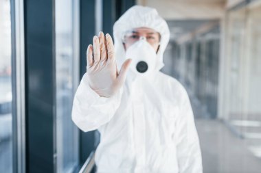 Stop the virus gesture. Female doctor scientist in lab coat, defensive eyewear and mask standing indoors.