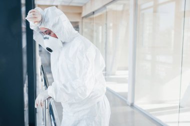 Feels bad and sick. Female doctor scientist in lab coat, defensive eyewear and mask standing indoors.