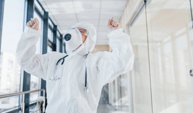 Zaferi kutluyoruz. Laboratuvar önlüklü, koruyucu gözlüklü ve maskeli kadın doktor içeride dikiliyor..
