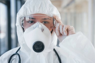 Laboratuvar önlüklü, koruyucu gözlüklü ve maskeli kadın doktor içeride dikiliyor..