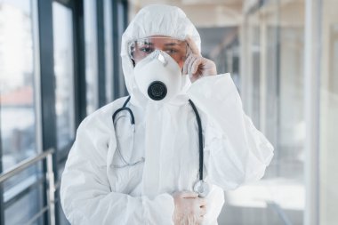 Laboratuvar önlüklü, koruyucu gözlüklü ve maskeli kadın doktor içeride dikiliyor..