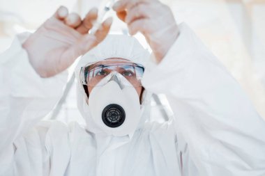 Şırınganın içinde ilaç var. Laboratuvar önlüklü, koruyucu gözlüklü ve maskeli kadın doktor içeride dikiliyor..