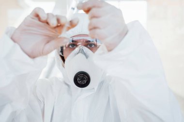 Şırınganın içinde ilaç var. Laboratuvar önlüklü, koruyucu gözlüklü ve maskeli kadın doktor içeride dikiliyor..