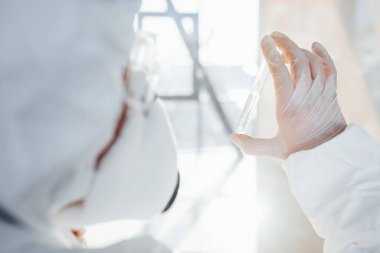 Elinde test tüpüyle. Laboratuvar önlüklü, koruyucu gözlüklü ve maskeli kadın doktor içeride dikiliyor..