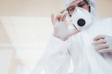 With test tube in hand. Female doctor scientist in lab coat, defensive eyewear and mask standing indoors.