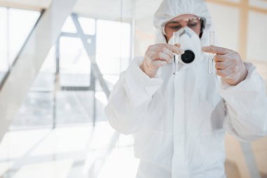 Elinde test tüpüyle. Laboratuvar önlüklü, koruyucu gözlüklü ve maskeli kadın doktor içeride dikiliyor..
