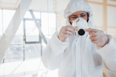 Elinde test tüpüyle. Laboratuvar önlüklü, koruyucu gözlüklü ve maskeli kadın doktor içeride dikiliyor..