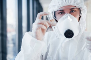 Elinde test tüpüyle. Laboratuvar önlüklü, koruyucu gözlüklü ve maskeli kadın doktor içeride dikiliyor..