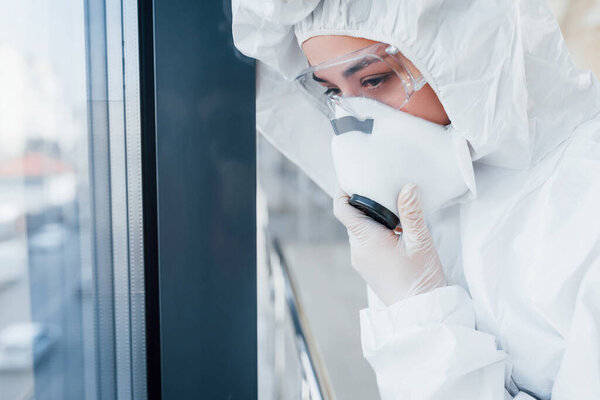 Feels bad and sick. Female doctor scientist in lab coat, defensive eyewear and mask standing indoors.