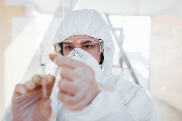 Holds syringe with medicine. Female doctor scientist in lab coat, defensive eyewear and mask standing indoors.