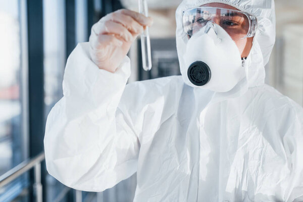 With test tube in hand. Female doctor scientist in lab coat, defensive eyewear and mask standing indoors.