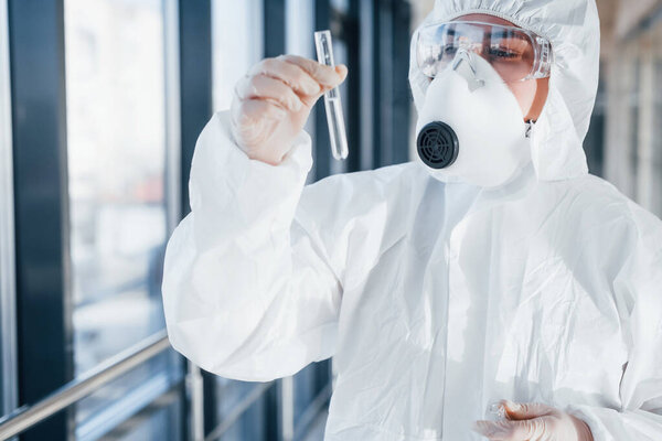 With test tube in hand. Female doctor scientist in lab coat, defensive eyewear and mask standing indoors.