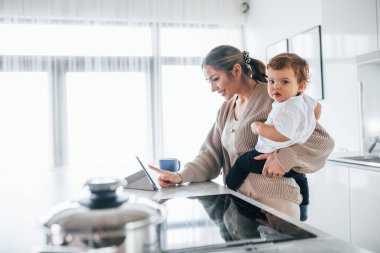 Female blogger mother with her kid together indoors at home at weekend using tablet.