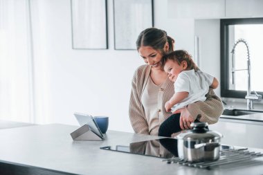 Female blogger mother with her kid together indoors at home at weekend using tablet.