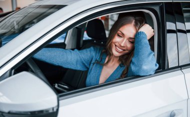 Positive woman in blue shirt sits inside of new brand new car. In auto salon or airport.