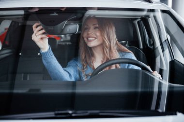 Front view of positive woman in blue shirt that sits inside of new brand new car. In auto salon or airport.