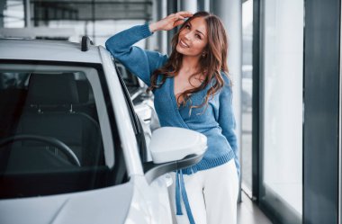 Mavi gömlekli olumlu bir kadın yepyeni bir arabaya yaslanıyor. Otomobil salonunda..