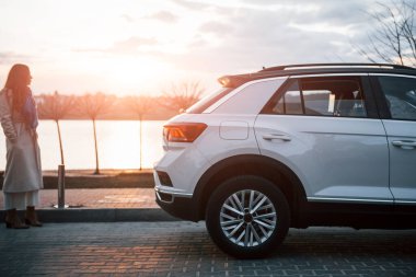 Sıcak beyaz elbiseli bir kadın beyaz modern arabanın yanında duruyor ve güneşin doğuşunu seyrediyor..